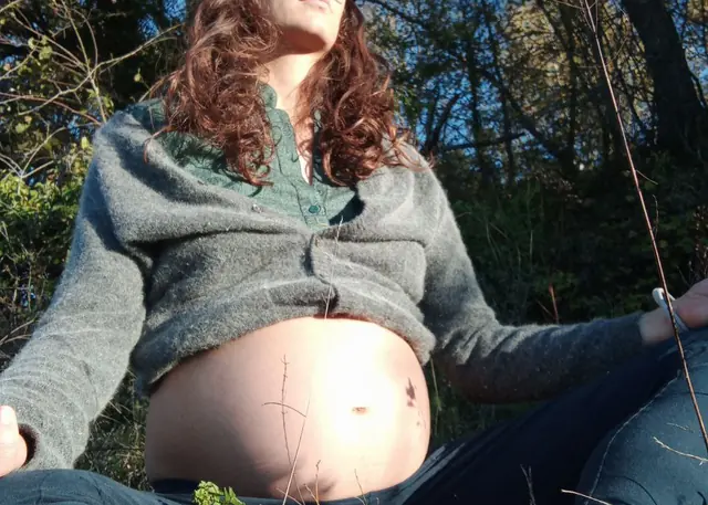 Eine schwangere Frau sitzt mit geschlossenen Augen im Sonnenlicht im Wald